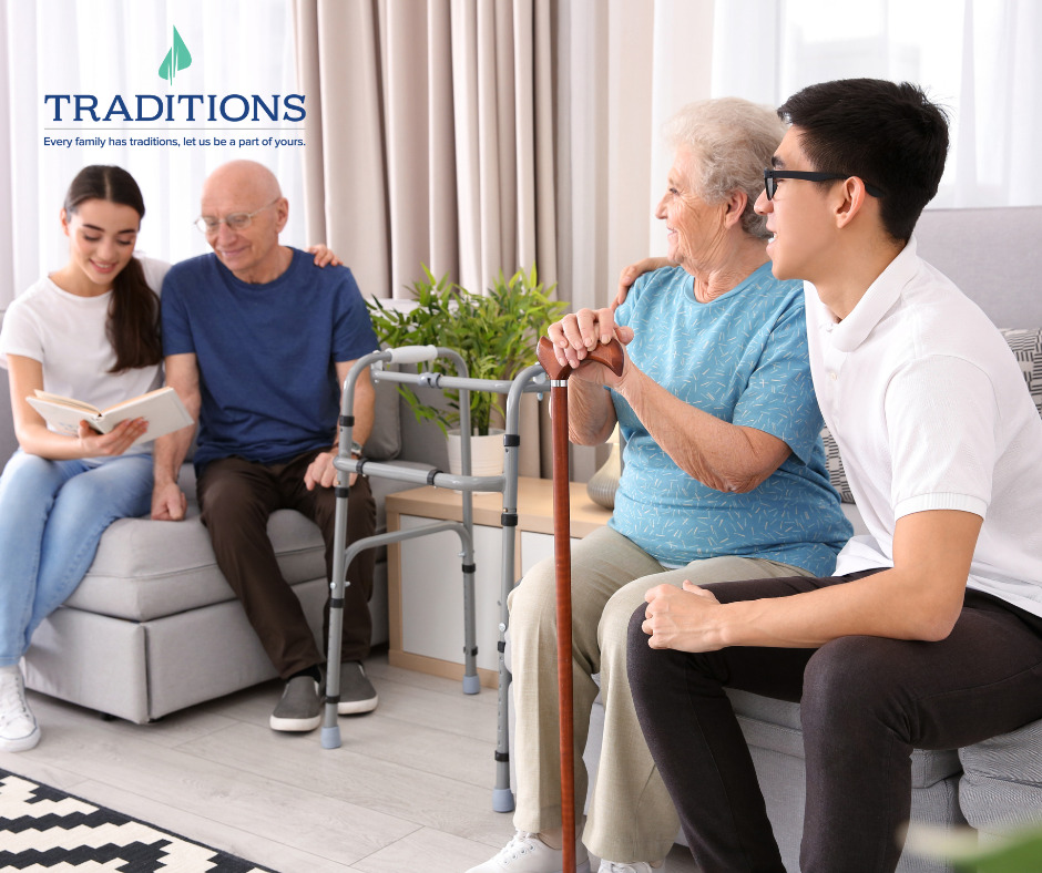 An elderly couple sitting with their grandchildren on the couch. A girl is sitting on the gray couch with her hand over her grandfathers shoulder and reading a book. A boy has his hands over his grandmothers shoulder and they are both looking at the girl reading. Traditions logo in the top left corner