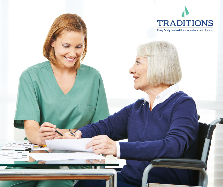 A caregiver and elderly woman sitting next to a table with crossword puzzles on it. Traditions logo on the top right corner