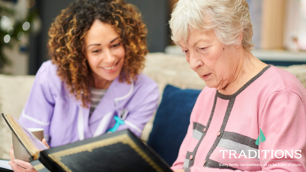 an elderly woman looks at a photo album next to a smiling young caregiver with Traditions logo on the bottom right corner