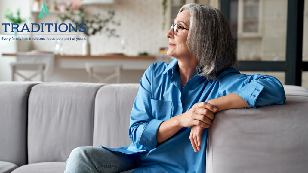 An elderly woman with glasses wearing a blue blouse and jeans sitting on a gray couch looking off in the distance. Traditions logo in the top left corner.