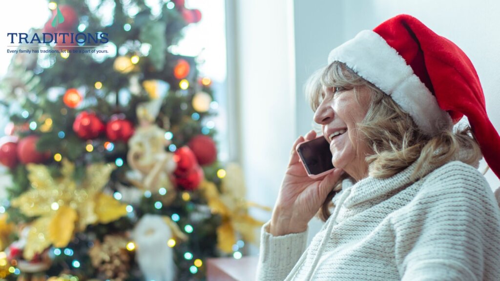 An elderly woman wearing a Santa hat talking on the phone with a Christmas tree in the background and Traditions logo at the top left corner.