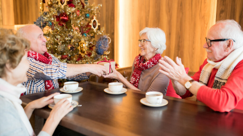4 Elderly People are sitting around a table with coffee, celebrating Christmas. A man is giving a woman a present with a red ribbon—decorated Christmas Tree in the background.