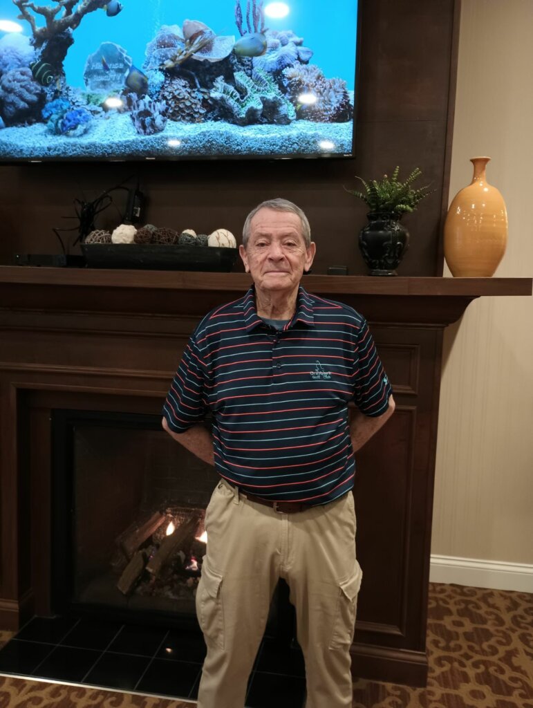 An elderly man standing in front of a fireplace with his hands behind his back while smiling
